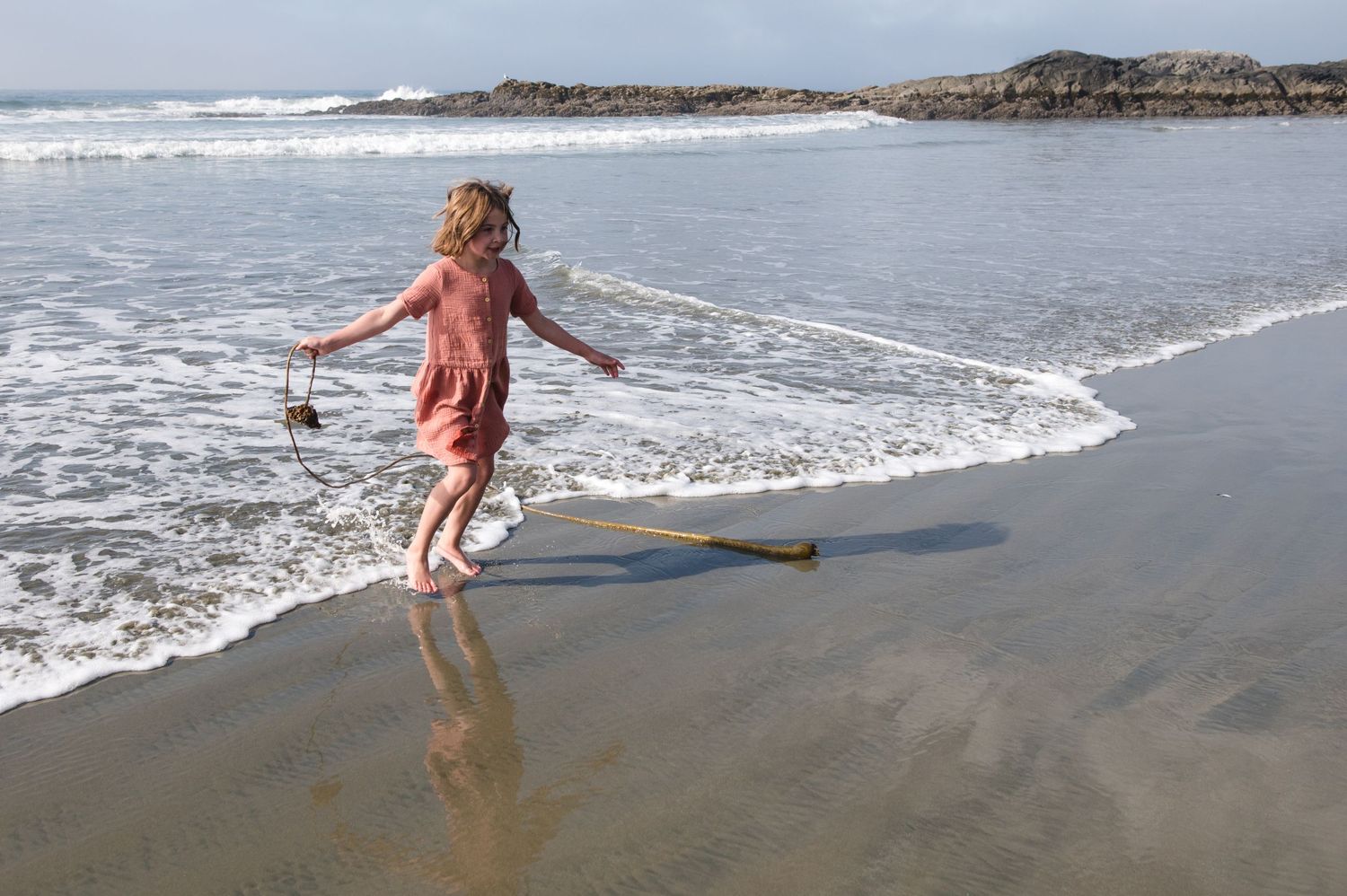 Child running through shallow ocean water with stick on sandy beach.