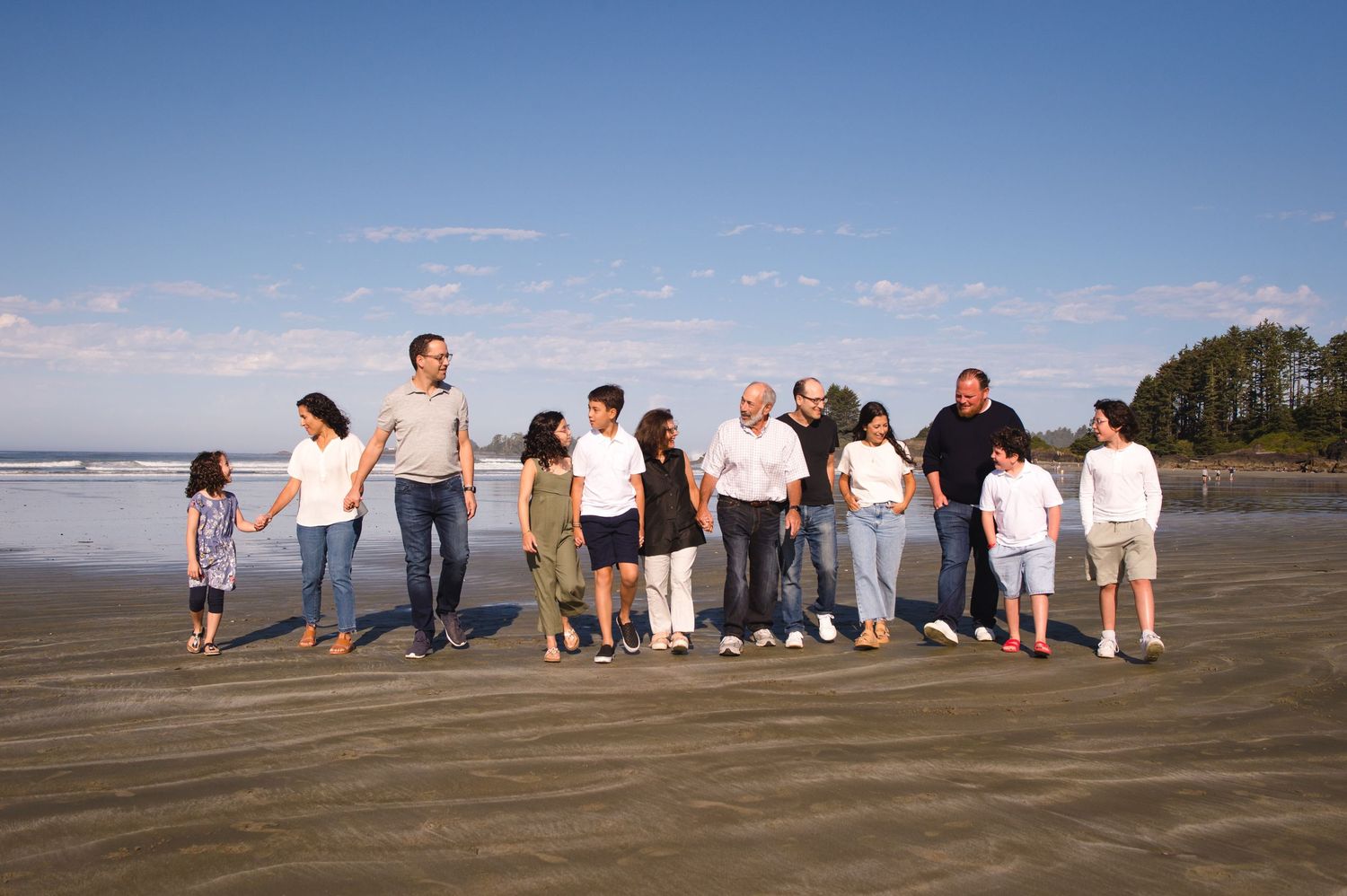 Extended family sitting together on sandy beach with ocean and blue sky in background.