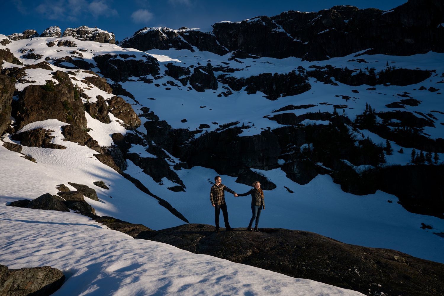 Two people holding hands standing on snow-covered rocky mountain terrain under dramatic blue sky.