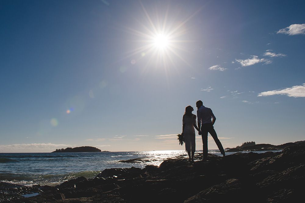 Silhouettes of people walking along a rocky coastline at sunset with bright sunburst in the blue sky.