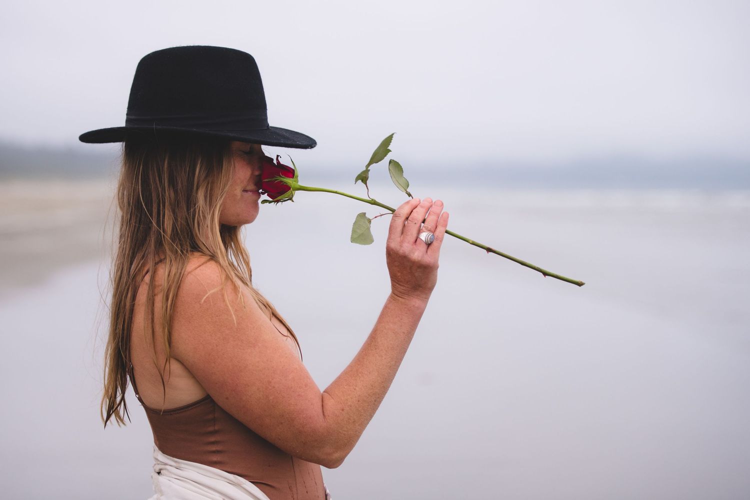 A woman in a brown swimsuit poses with a rose on a cloudy beach.