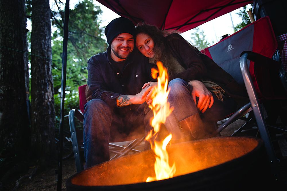 A group gathers around a blazing campfire in the woods at dusk under a red umbrella.