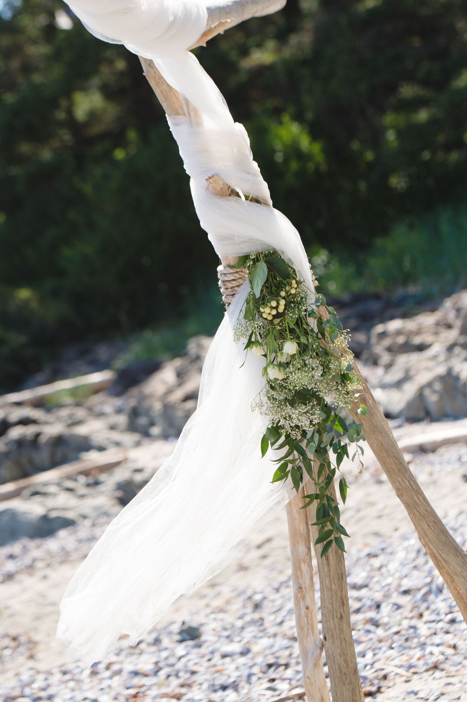 White fabric wedding arch decorated with greenery and flowers on a rocky beach.