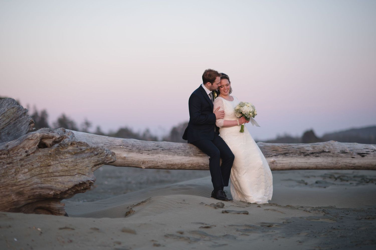Newlyweds share a tender moment while sitting on driftwood at dusk.