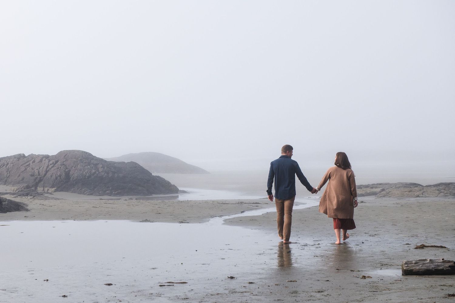 A couple walks hand in hand on a misty beach with mountains in the background.
