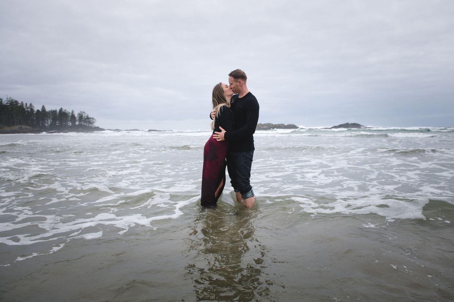 A romantic embrace in the shallow ocean waves with evergreen trees visible along the coastline.