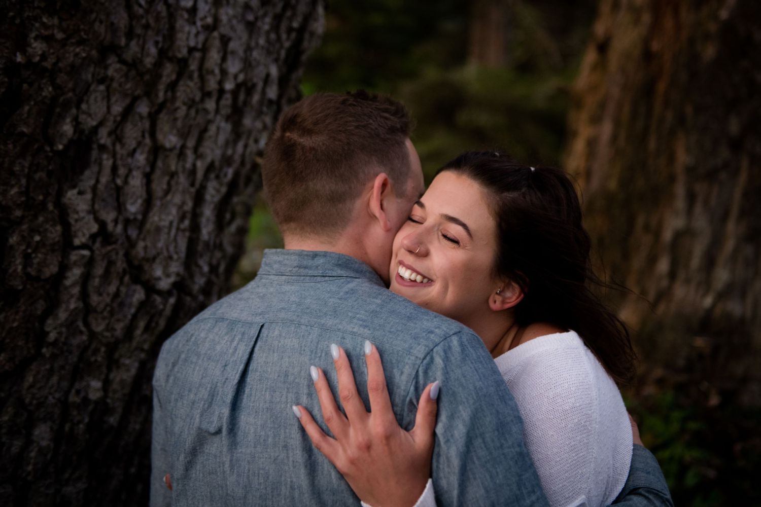 Two people share a tender embrace against a tree in a forest setting.