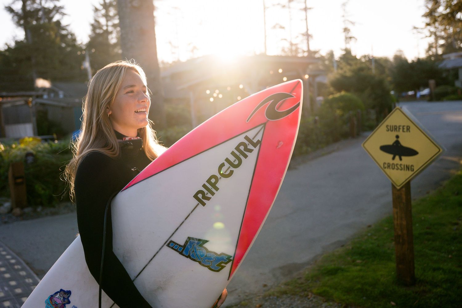 Young blonde woman in wetsuit holding a Rip Curl surfboard outdoors at golden hour near a surfer crossing sign.