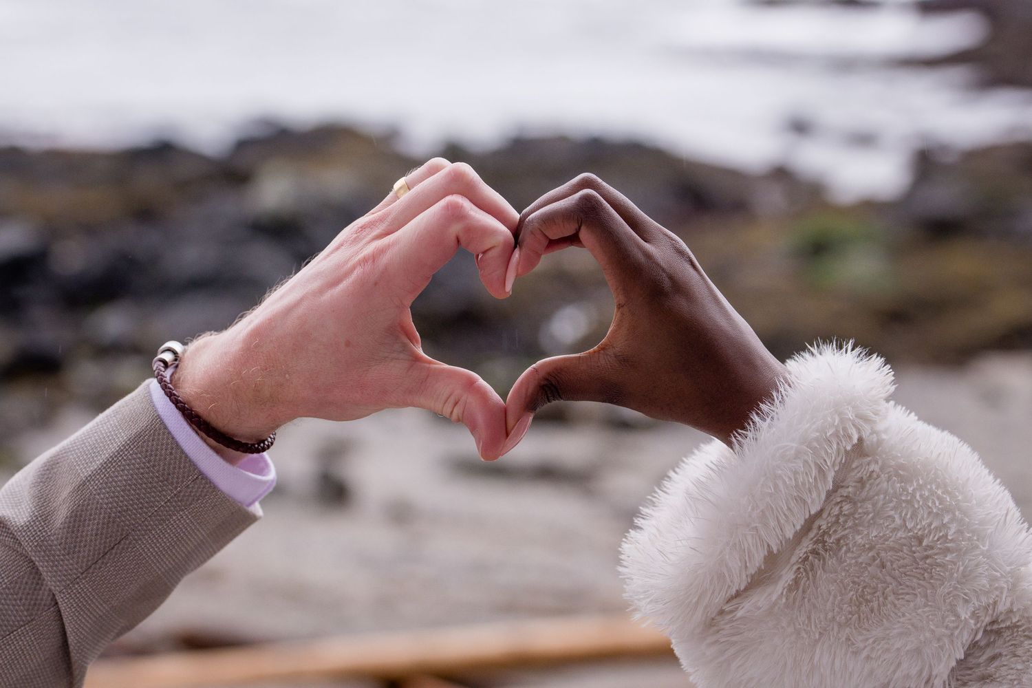 Two hands form a heart shape against a blurred coastal background.