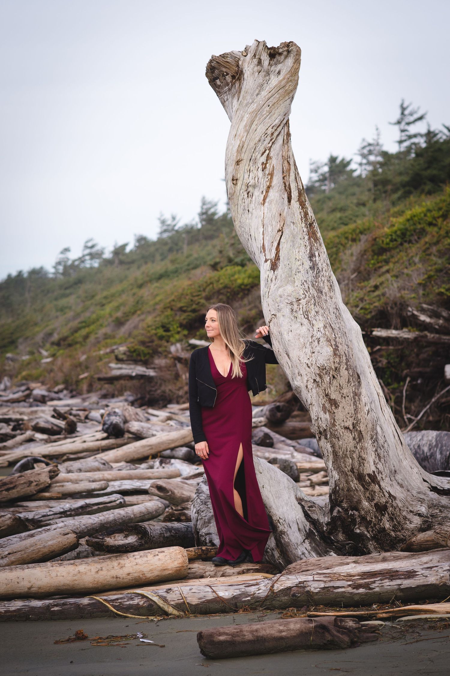 Person in burgundy dress sitting among driftwood on a Pacific Northwest beach.