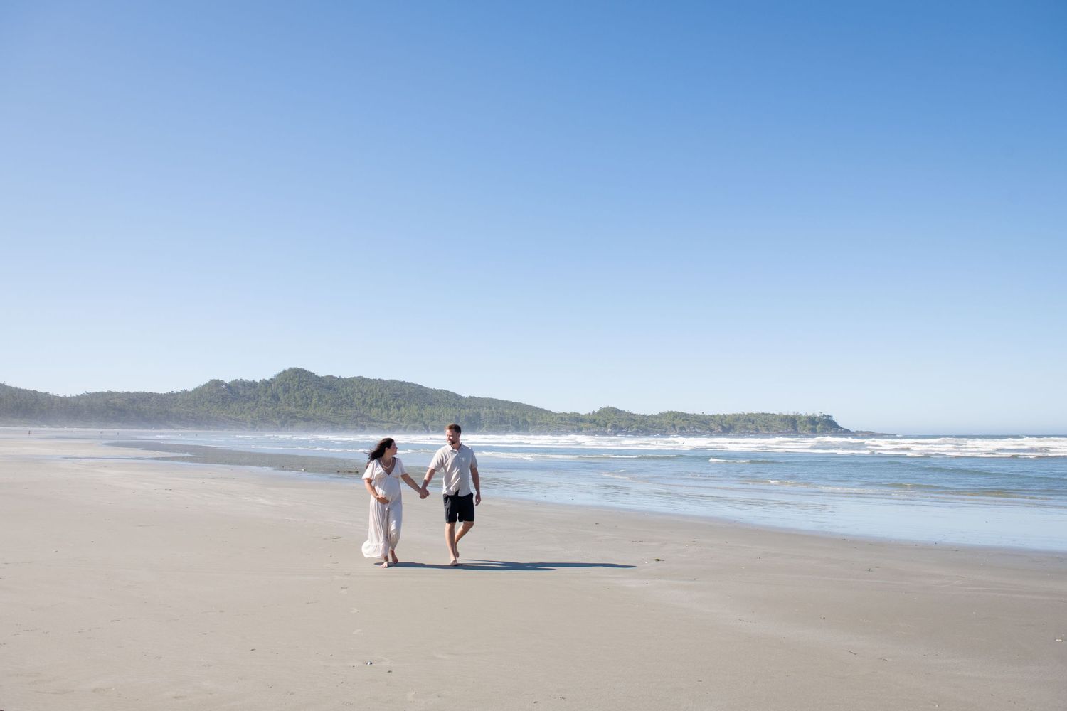 Two people walking along an empty beach with mountains visible in the distance.