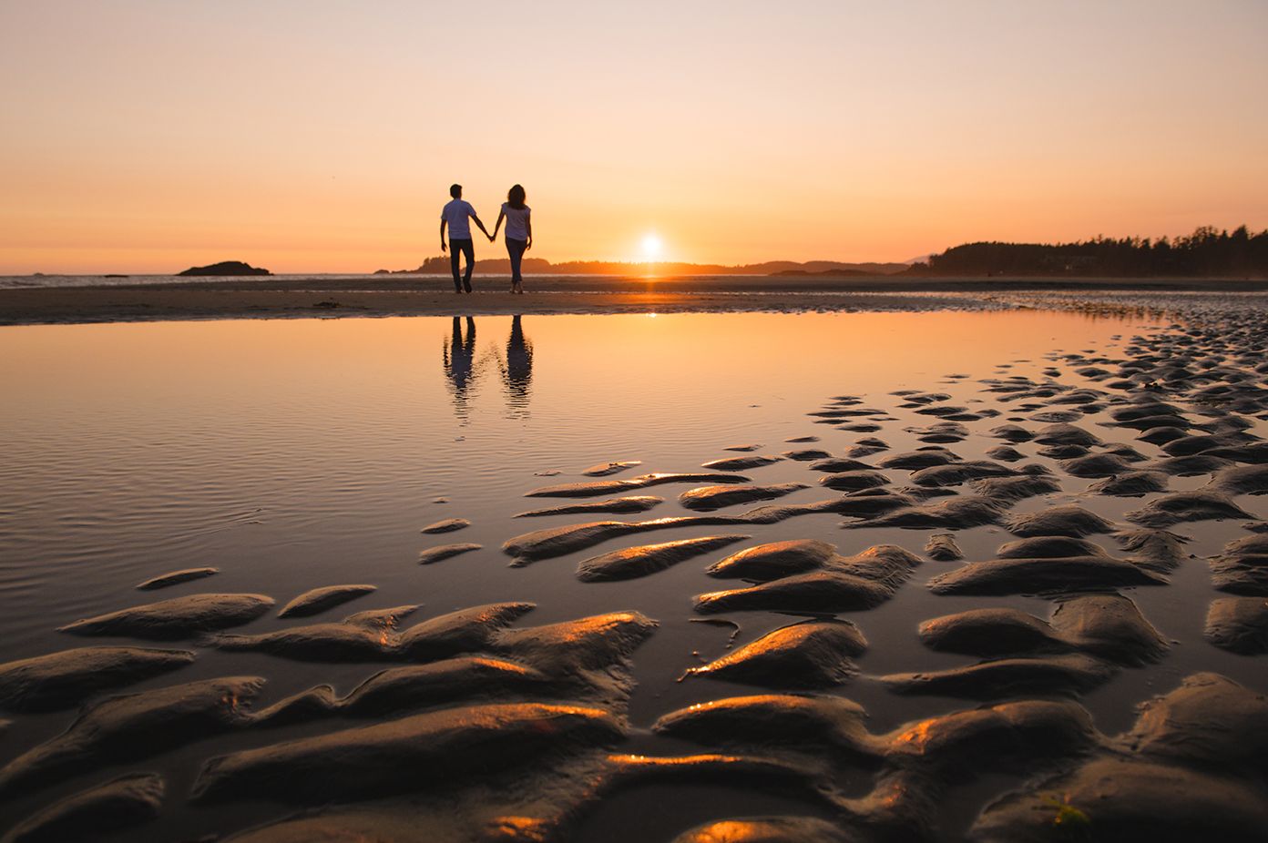 Two silhouettes walk along a sandy beach at sunset with rippled sand patterns and golden light reflecting on the water.