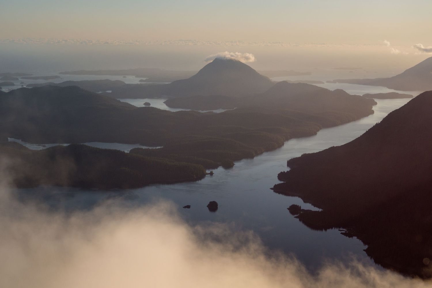 Aerial view of misty fjords and volcanic mountains reflected in calm waters at golden hour in Tofino and Clayoquot Sound.