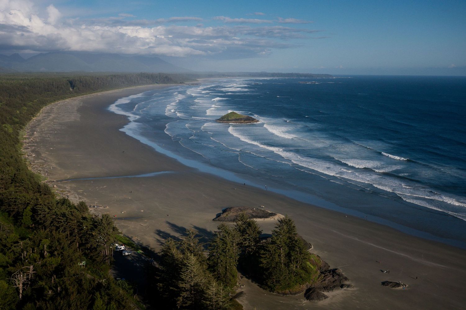 Aerial view of Long Beach in Tofino, a sandy beach with dark blue ocean waves, lush forest edge, and a rocky outcrop under cloudy skies.
