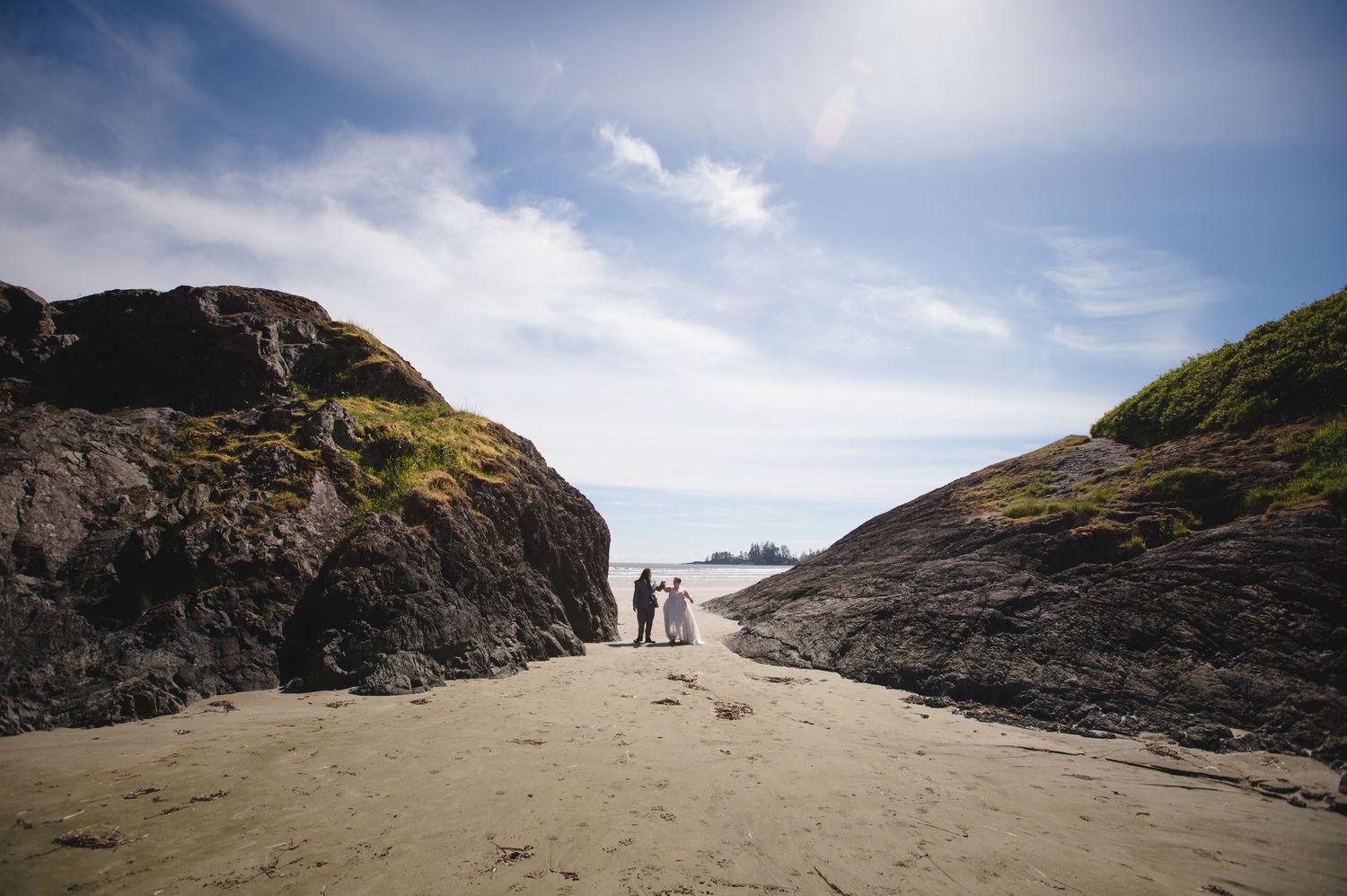 Two figures stand between rocky cliffs on a secluded beach under bright blue skies.