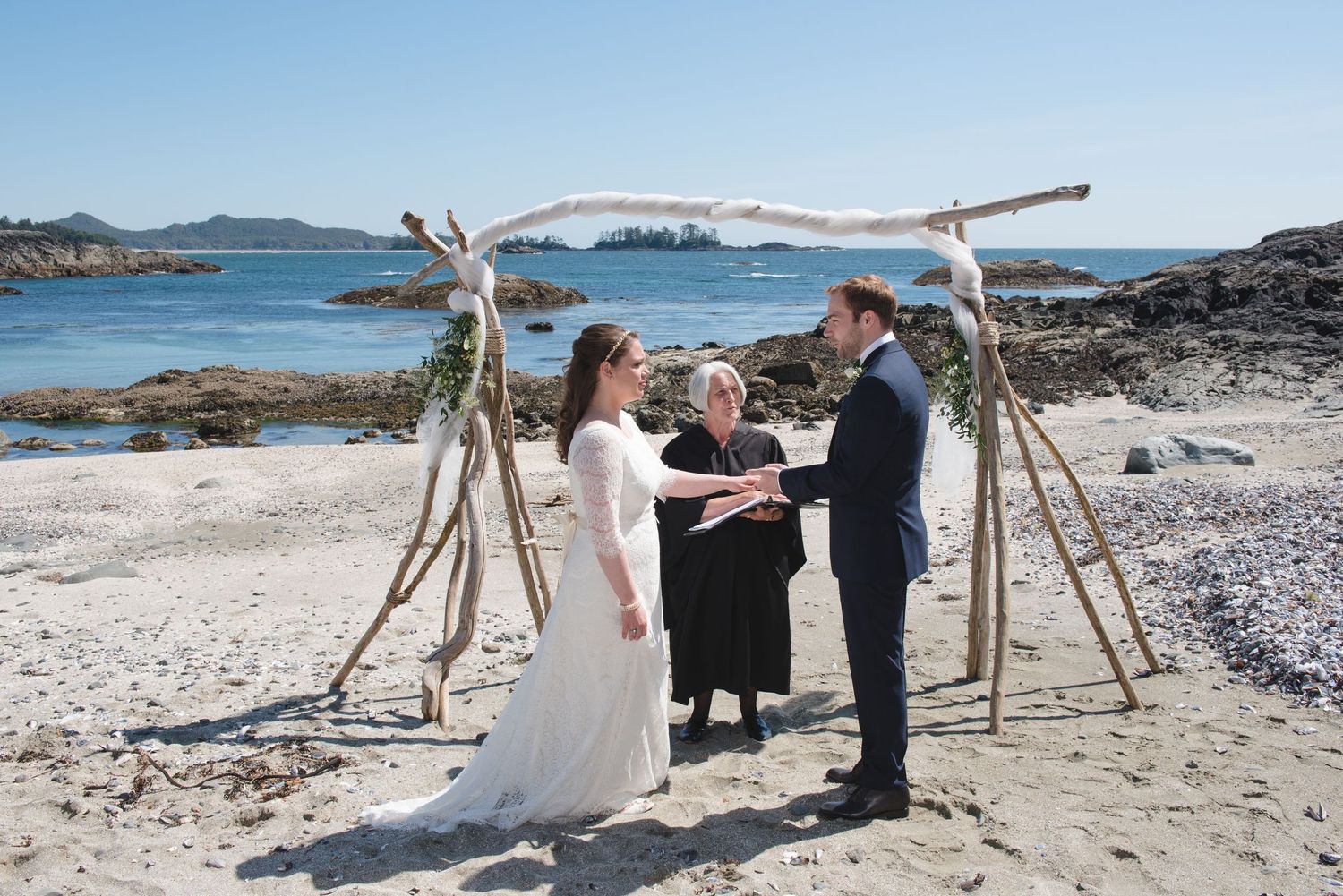 Beach wedding ceremony setup with driftwood arch and chairs on rocky shoreline.