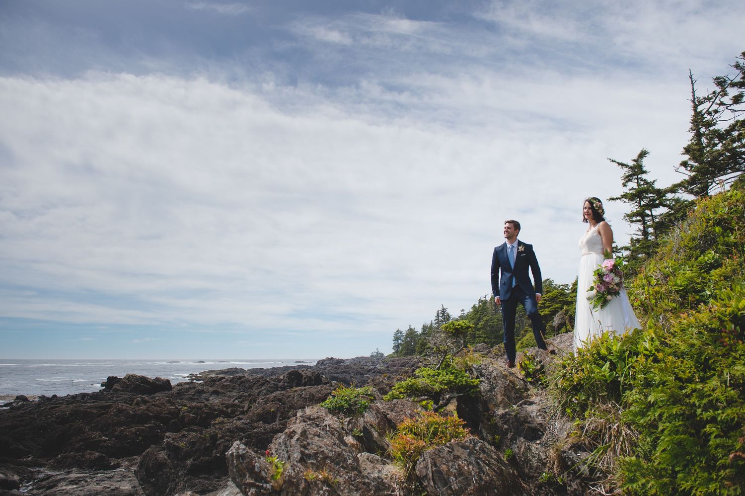 A couple stands on rocky coastal cliffs overlooking the Pacific Ocean on their wedding day.