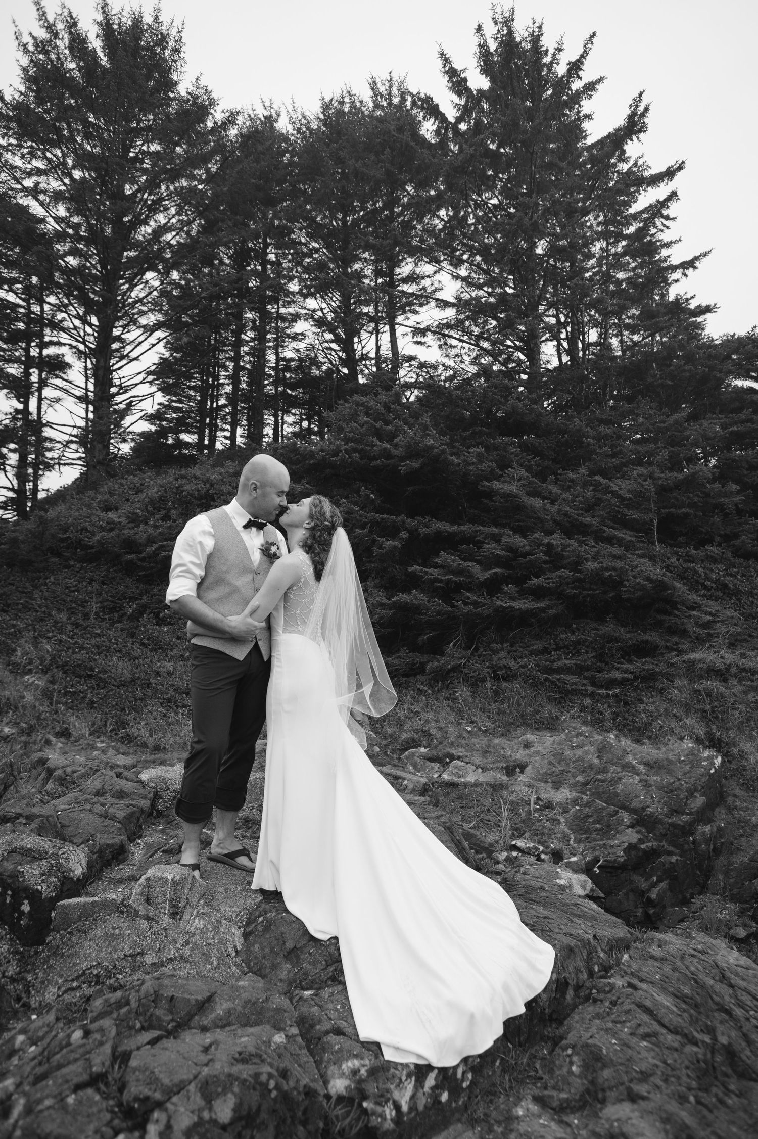 A romantic black and white wedding photo captured against a backdrop of tall pine trees on rocky terrain.