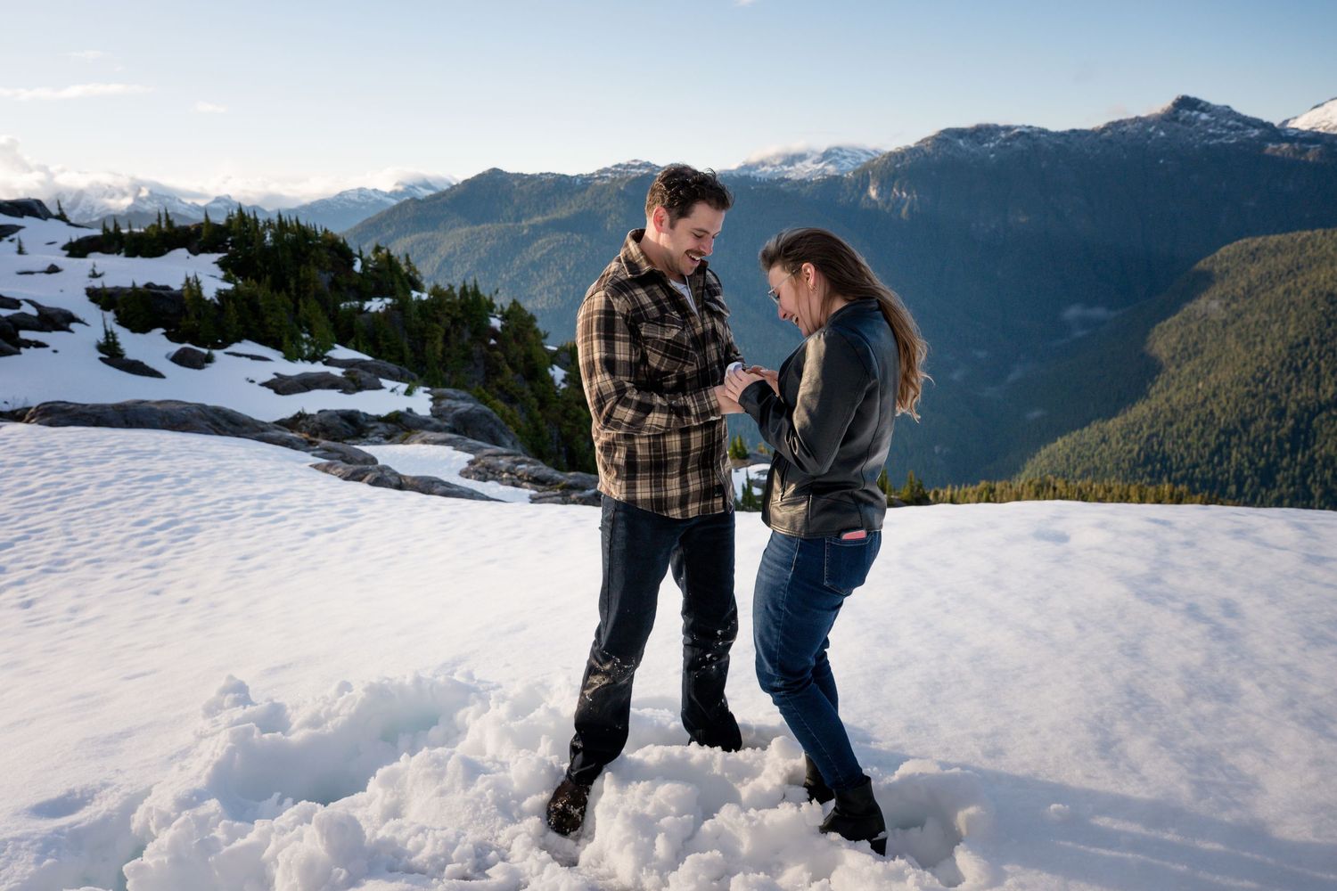 Couple holding hands on a snowy mountain peak with scenic evergreen forest and blue sky backdrop.