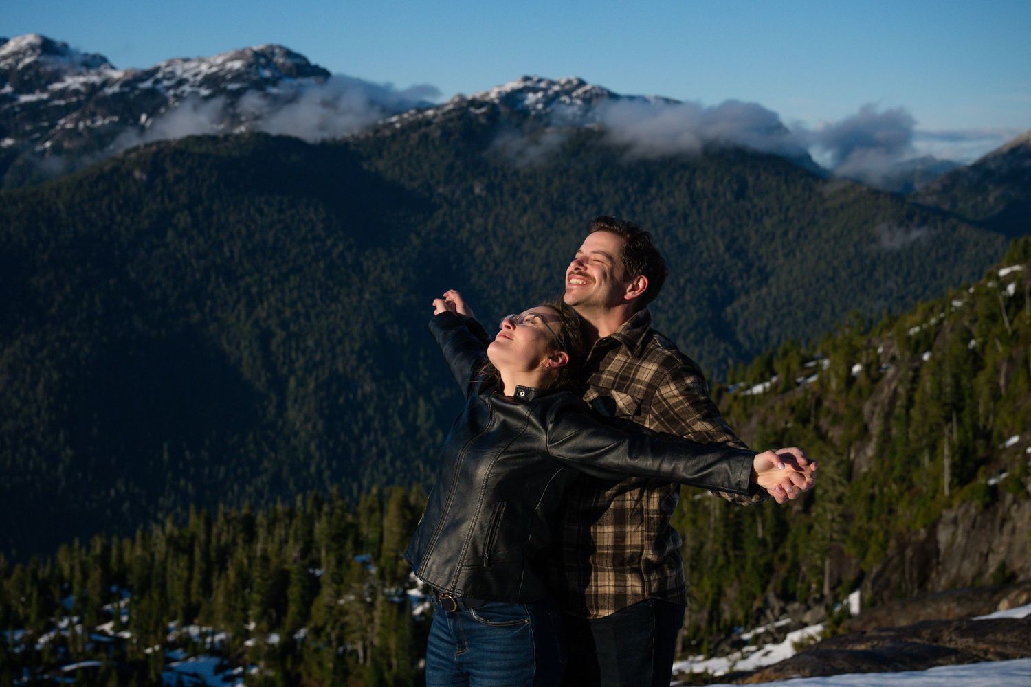Couple embracing with arms outstretched enjoying panoramic mountain views on a sunny day.