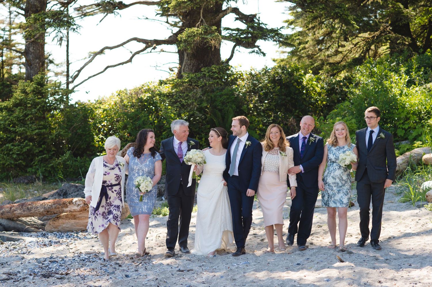 Wedding group photo with guests lined up along rocky beach with pine trees in background.