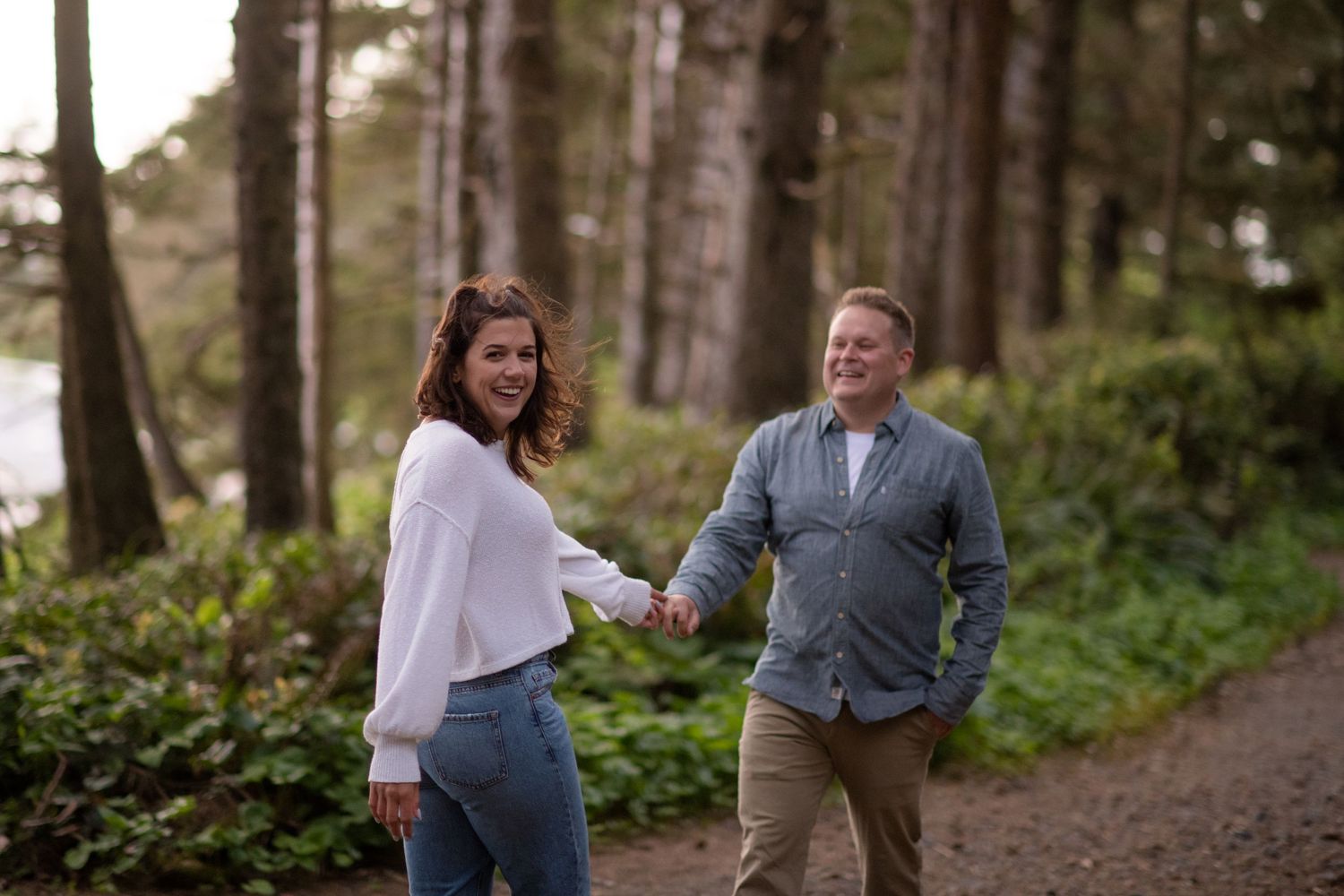 A couple walks hand in hand along a forest trail surrounded by tall trees.