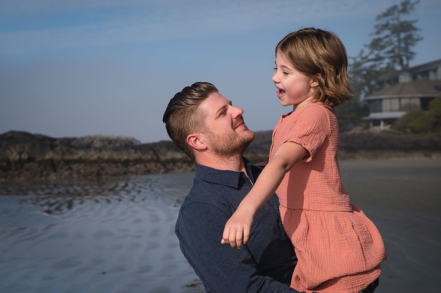 A couple shares tender moments on a scenic beach with driftwood and mountains in the background.