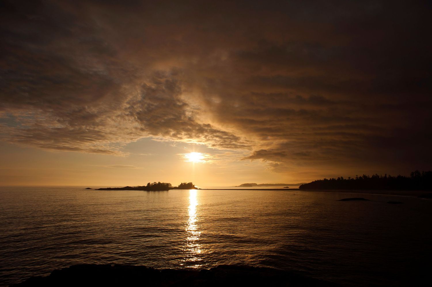 Dramatic sunset over calm ocean waters with silhouetted islands and golden rays reflecting on the water's surface.