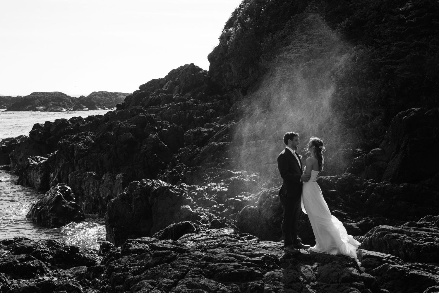 A dramatic black and white wedding photo on rocky coastal cliffs with ocean waves in the background.