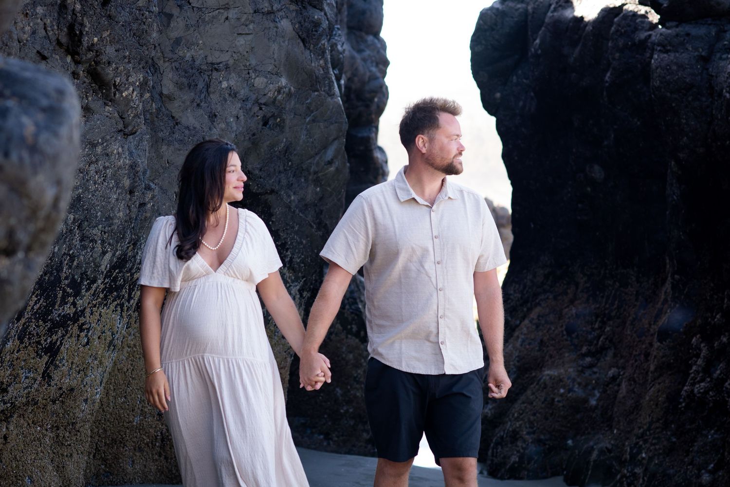 A romantic couple poses against dark rocky cliffs near the ocean during a photo session.