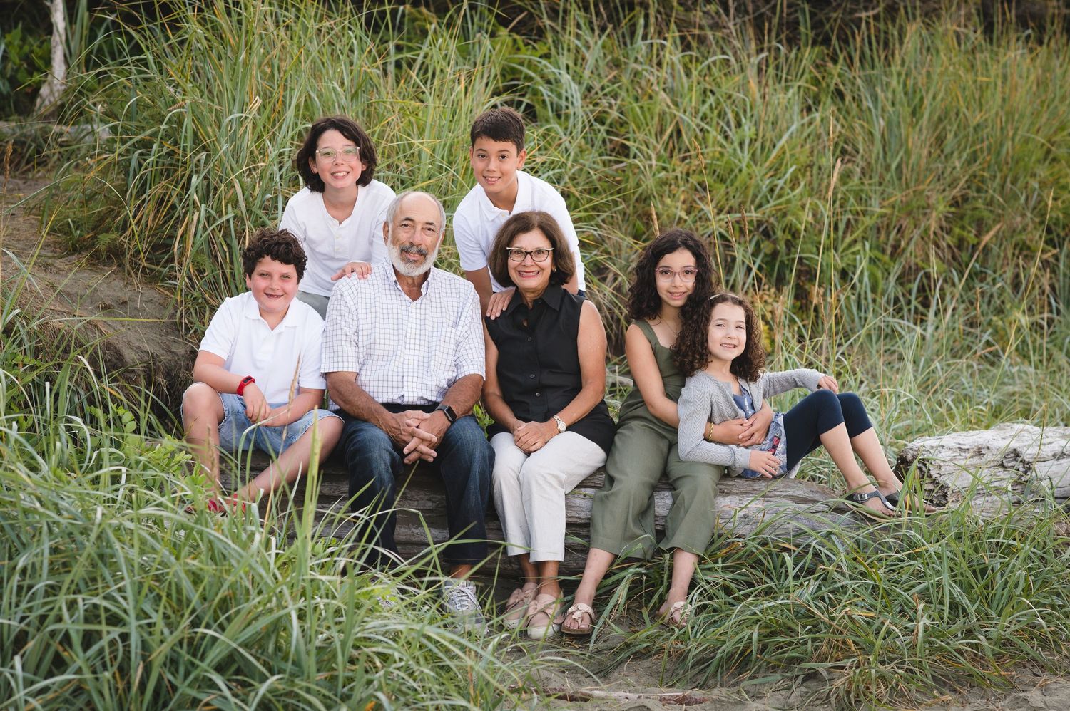 A multi-generational family group poses together on rocky steps surrounded by tall beach grass.