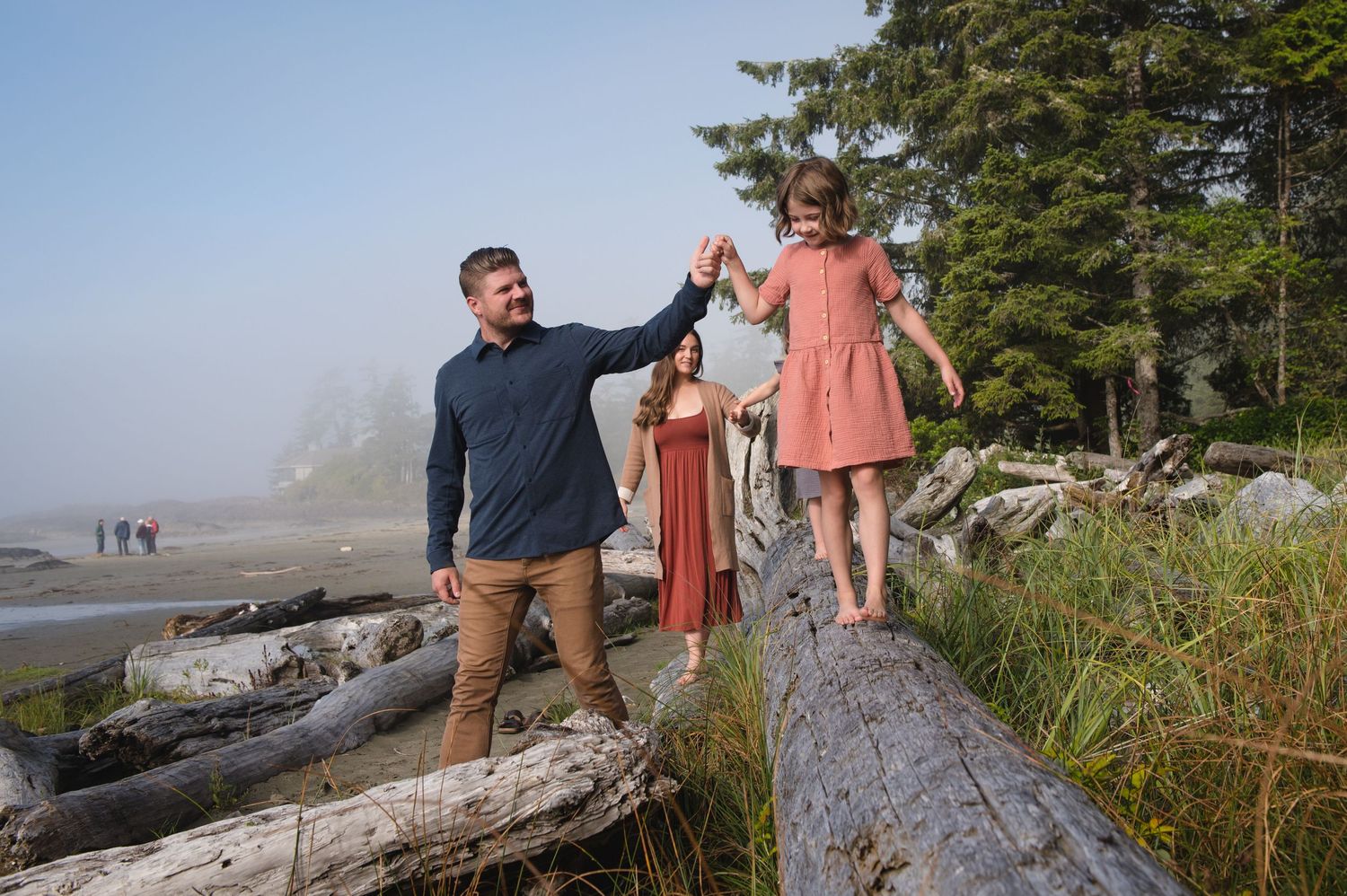 Family photo session on a coastal beach with large driftwood logs and evergreen trees.