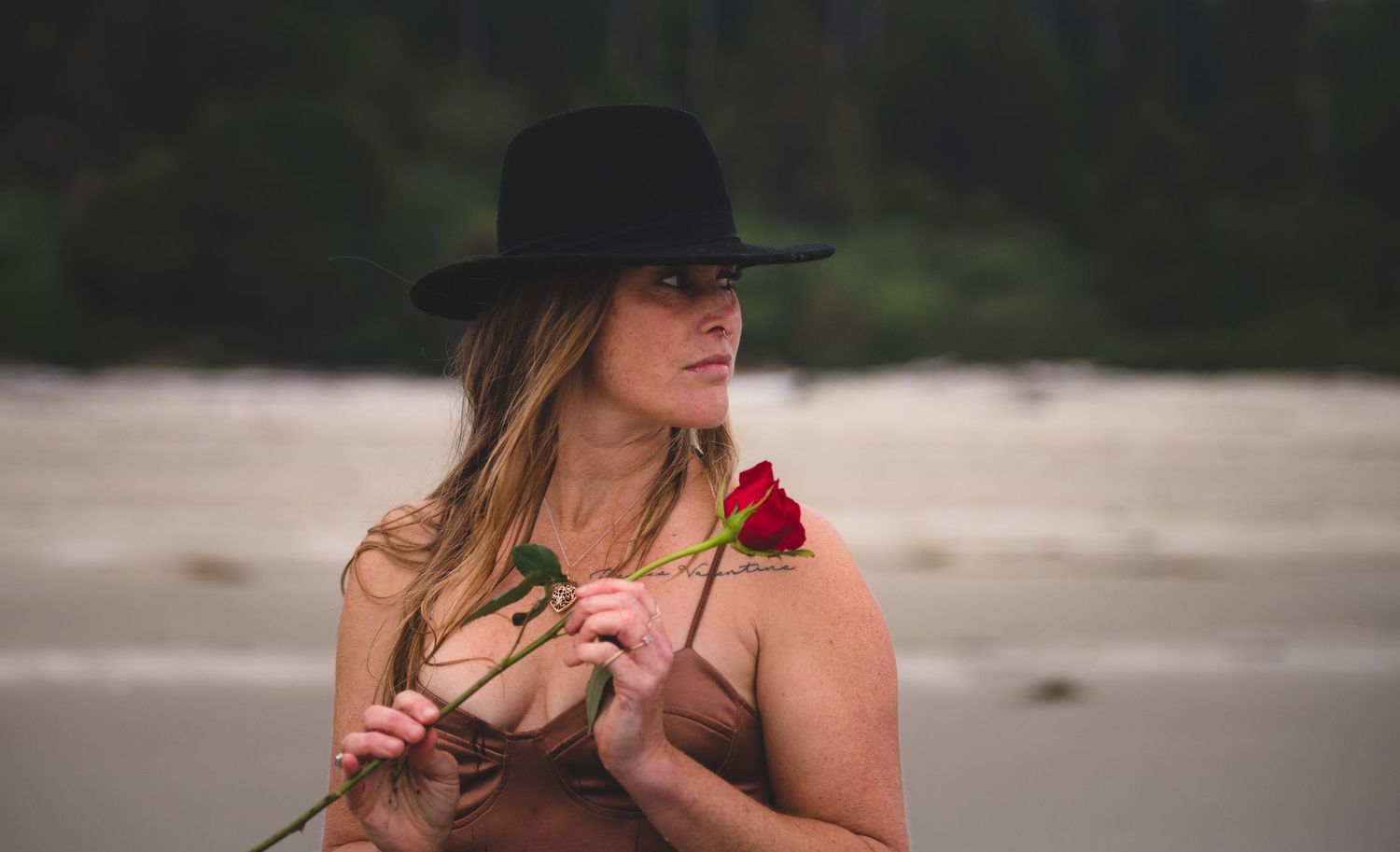 A person in a black hat poses on a beach with a red rose during a cloudy day.