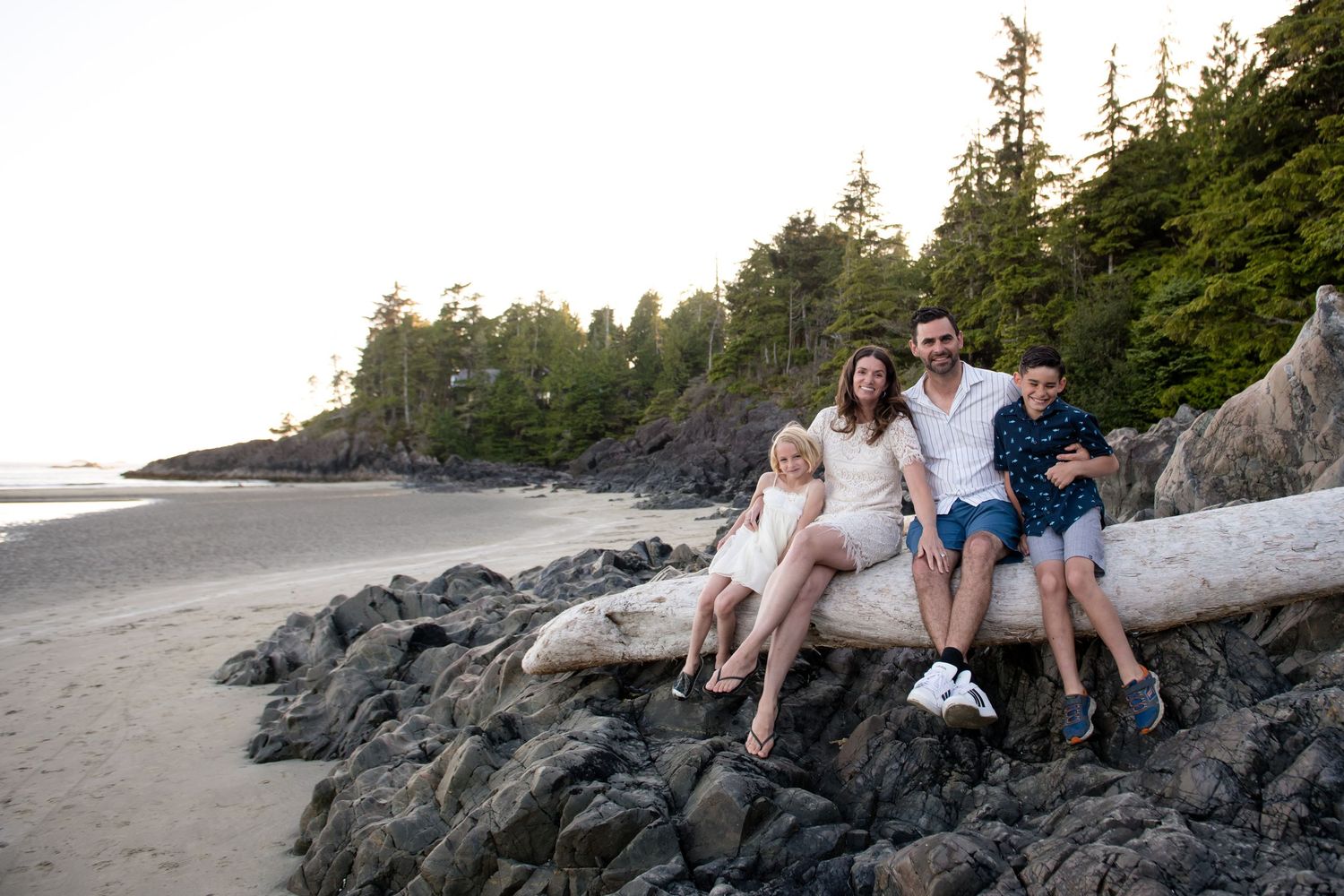 Three people sit together on driftwood along a rocky Pacific Northwest beach at sunset.