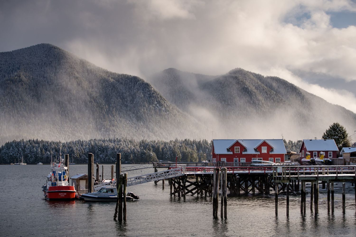 Misty coastal landscape with wooden buildings and docks along the shoreline beneath dramatic mountain peaks.