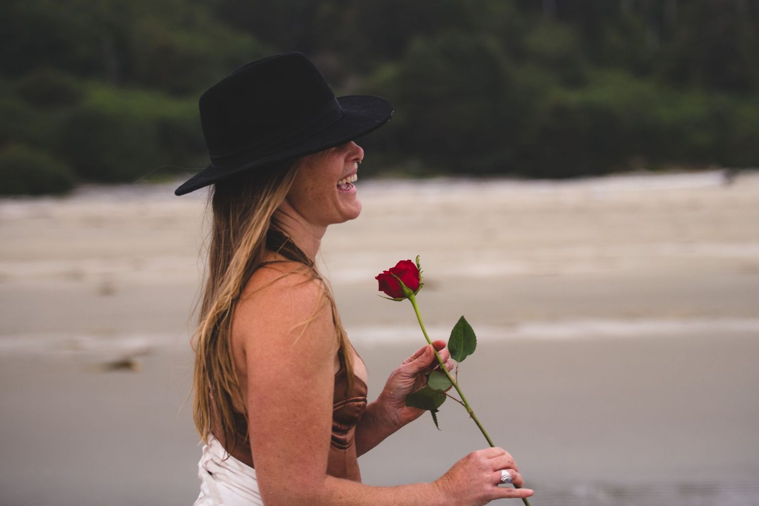 Person holding red rose walks on sandy beach with dark trees in background.