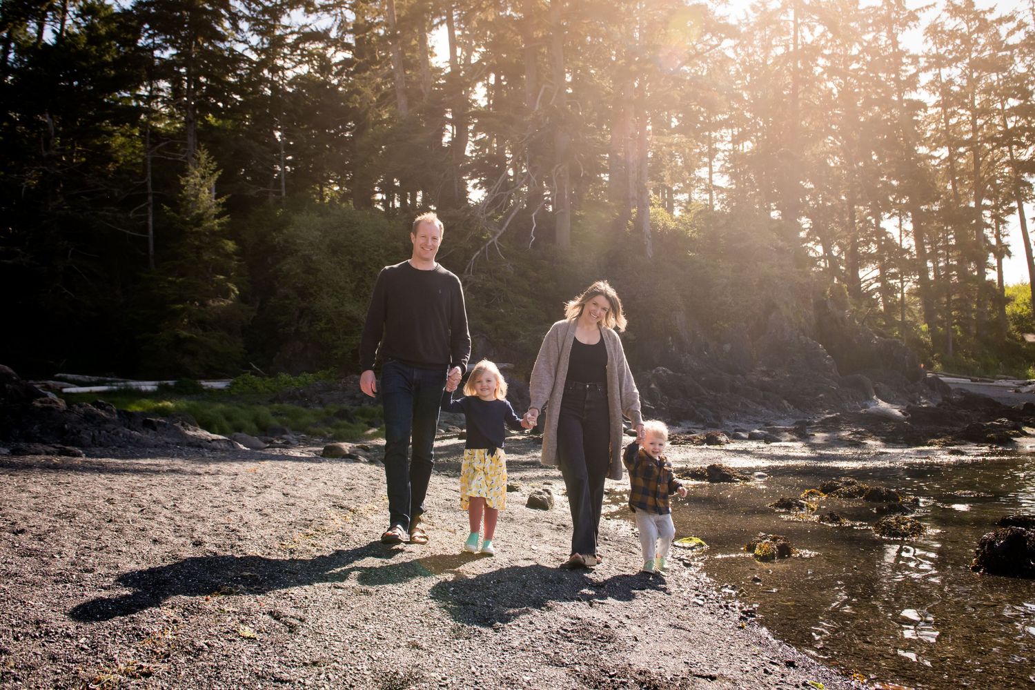 A family walks along a rocky beach at sunset with golden light filtering through evergreen trees in the background.