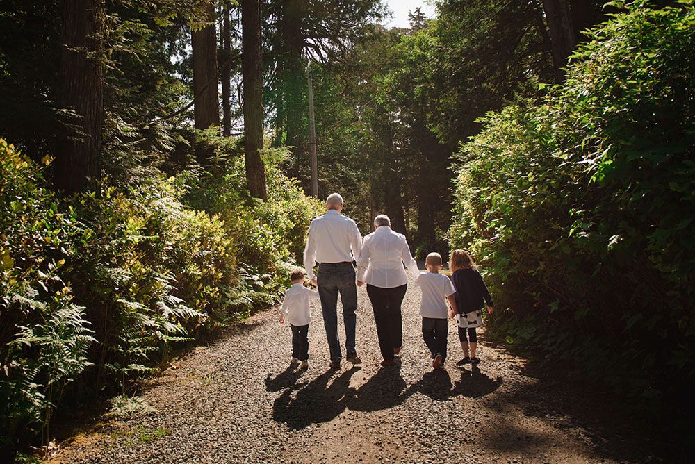 A family walks together on a sunlit forest path surrounded by lush greenery and tall trees casting long shadows.