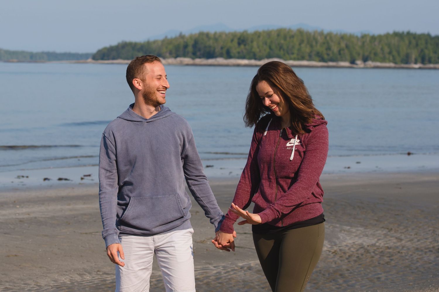 Couple walking hand in hand along a scenic beach with ocean and forested coastline in the background.
