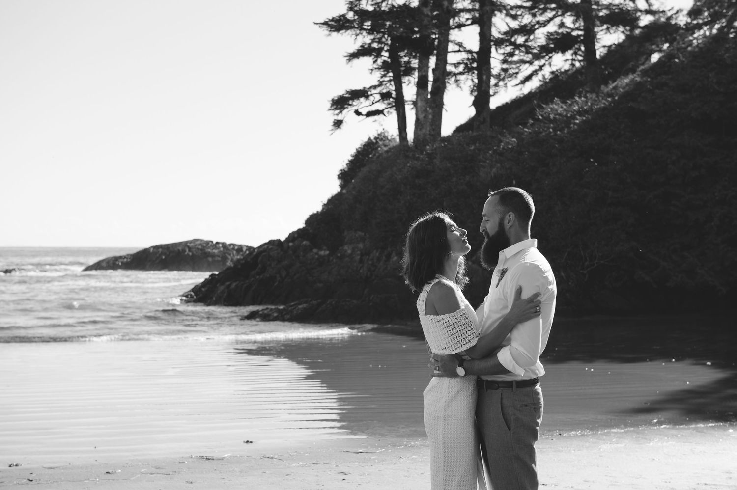 Couple in white embraces on a rocky beach during sunset with waves crashing in the background.