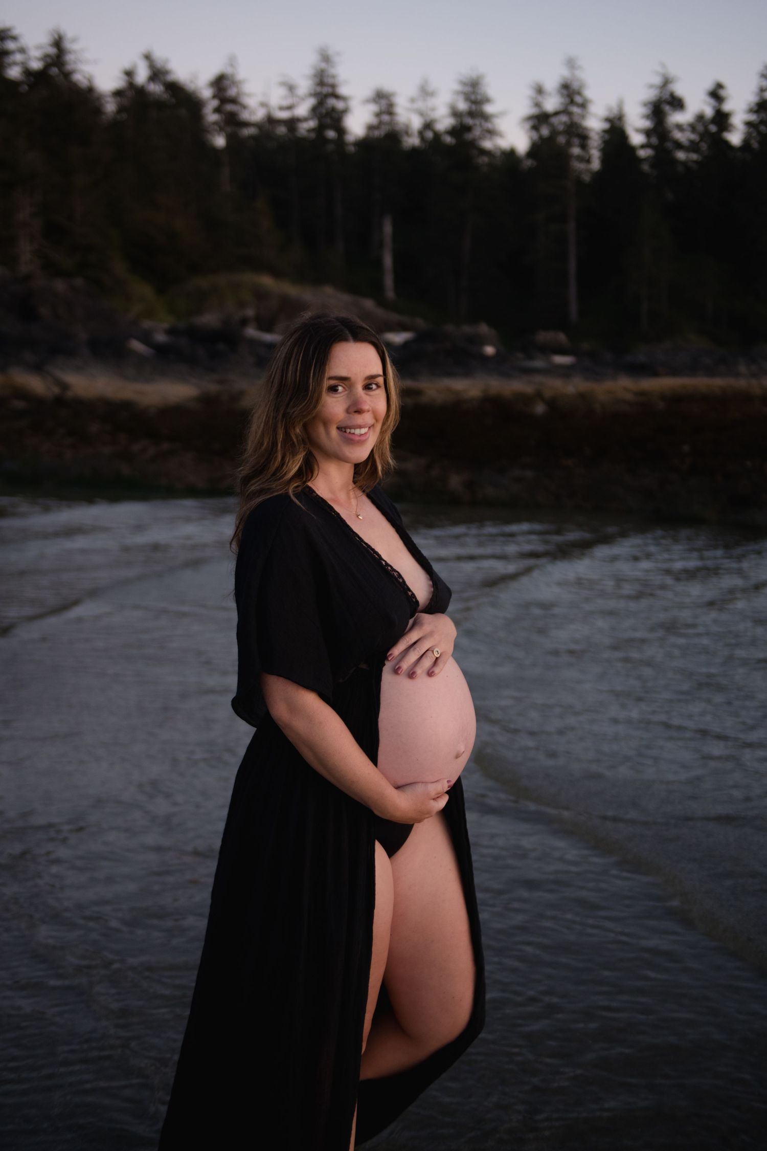 Maternity photo session at dusk by a serene waterfront with evergreen trees in the background.