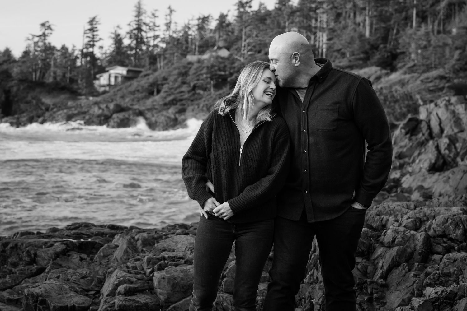 A romantic black and white moment captured on rocky coastal cliffs with evergreen trees in the background.
