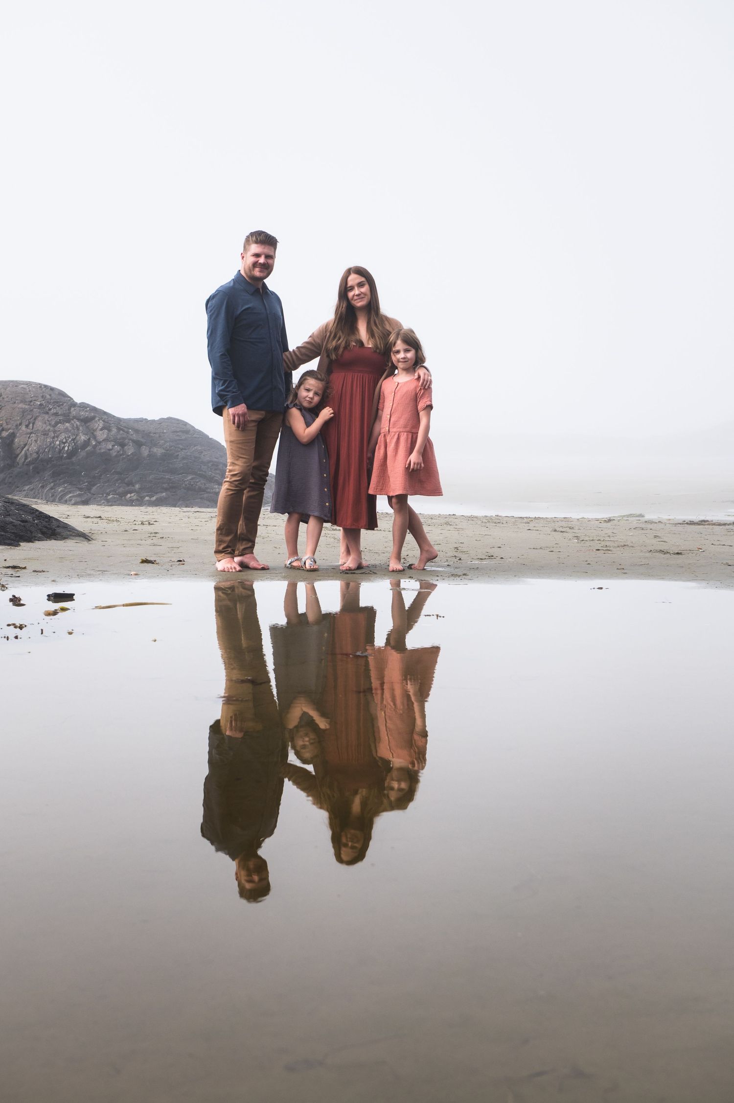 Family walking along misty beach with mountain backdrop reflected in wet sand at low tide.