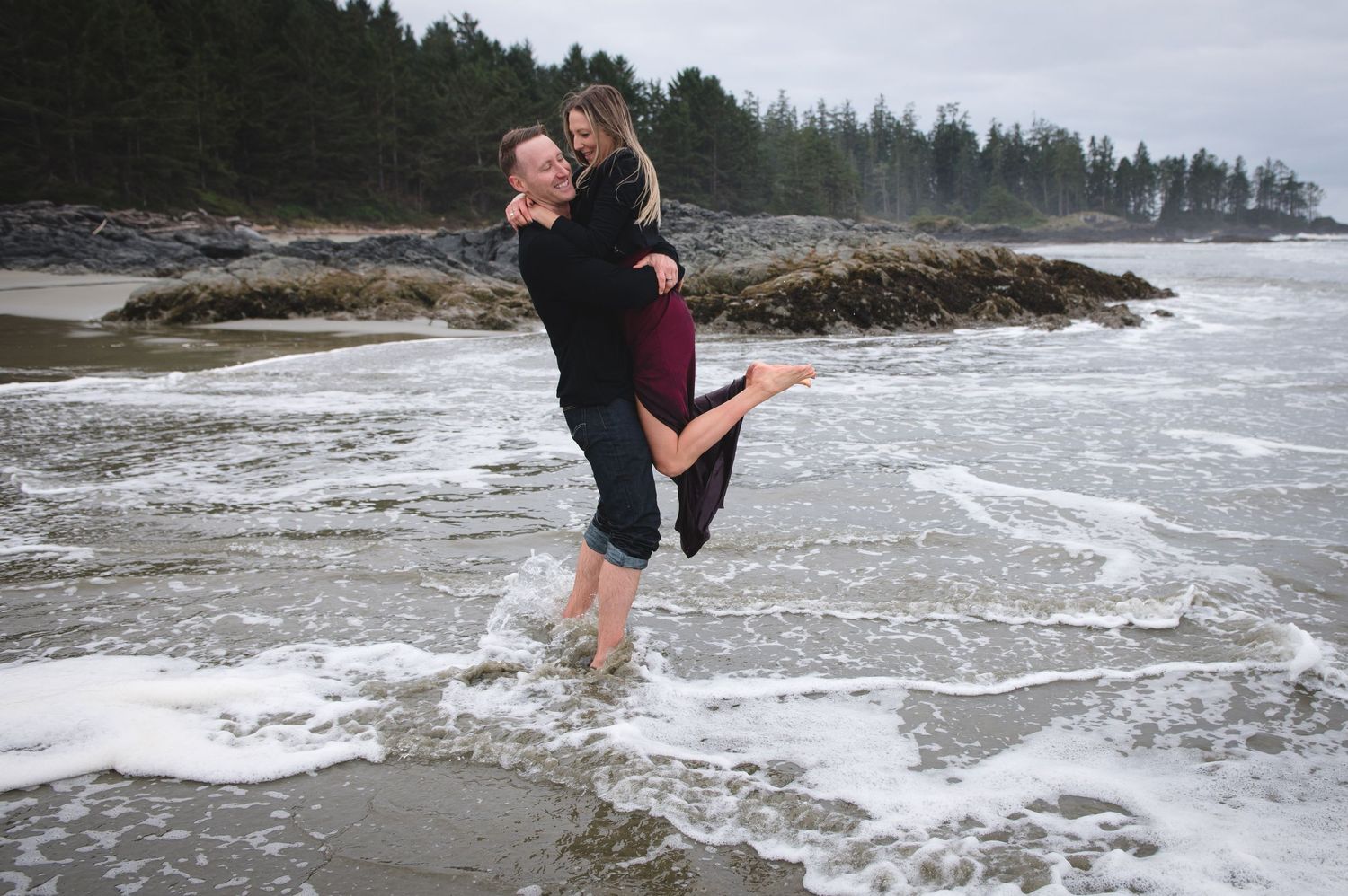 A joyful moment captured as a couple embraces in the ocean surf on a rocky beach.