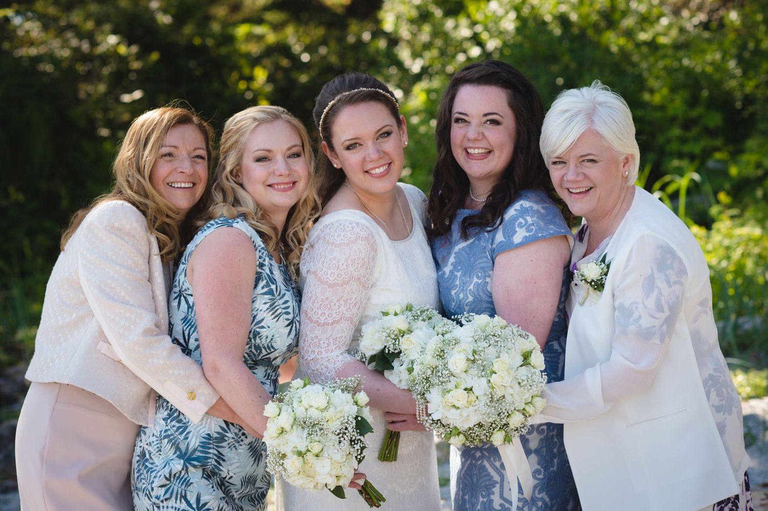 Wedding party gathered together holding white flower bouquets on sunny beach.
