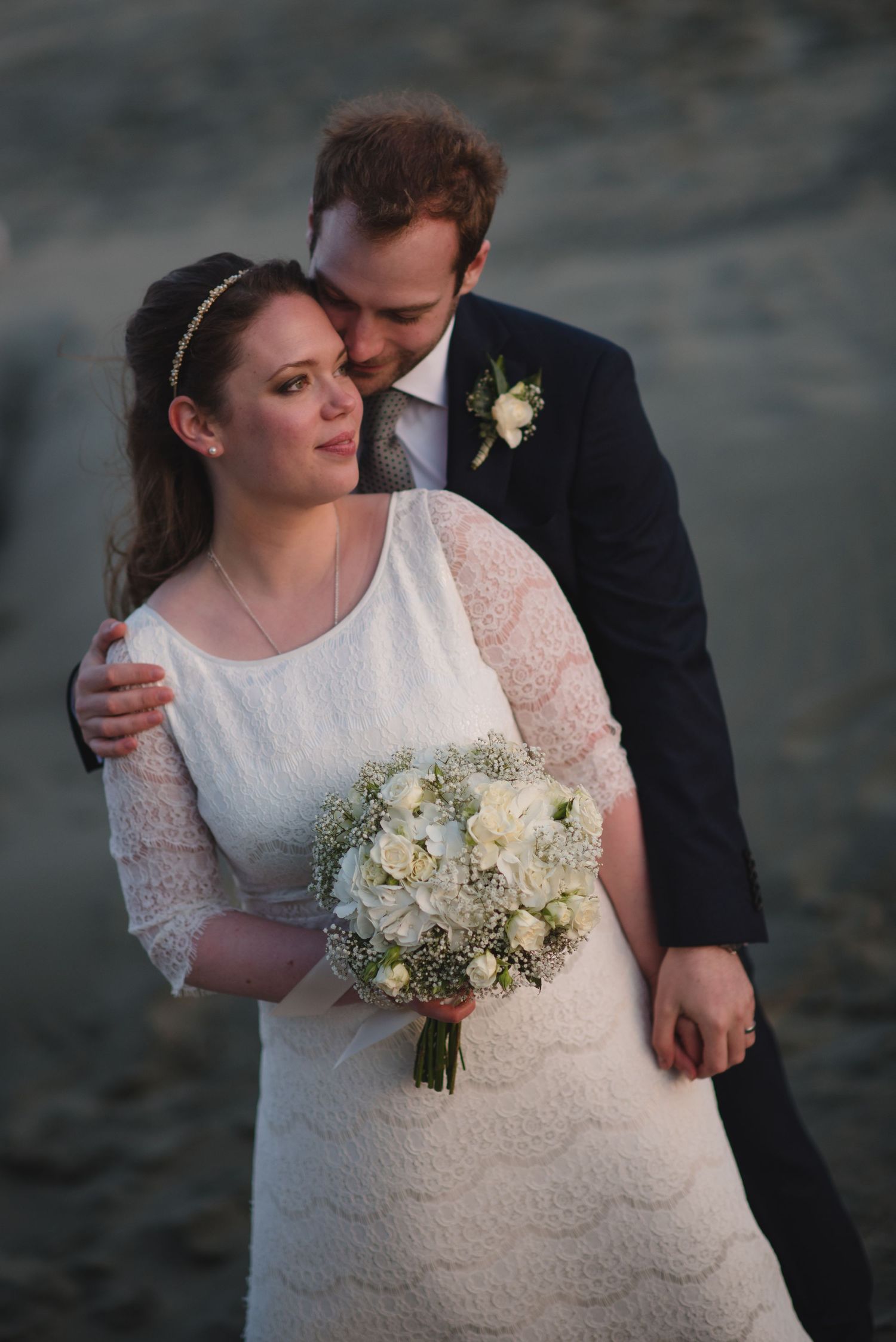 A tender wedding portrait with white bouquet against dusky coastal background.