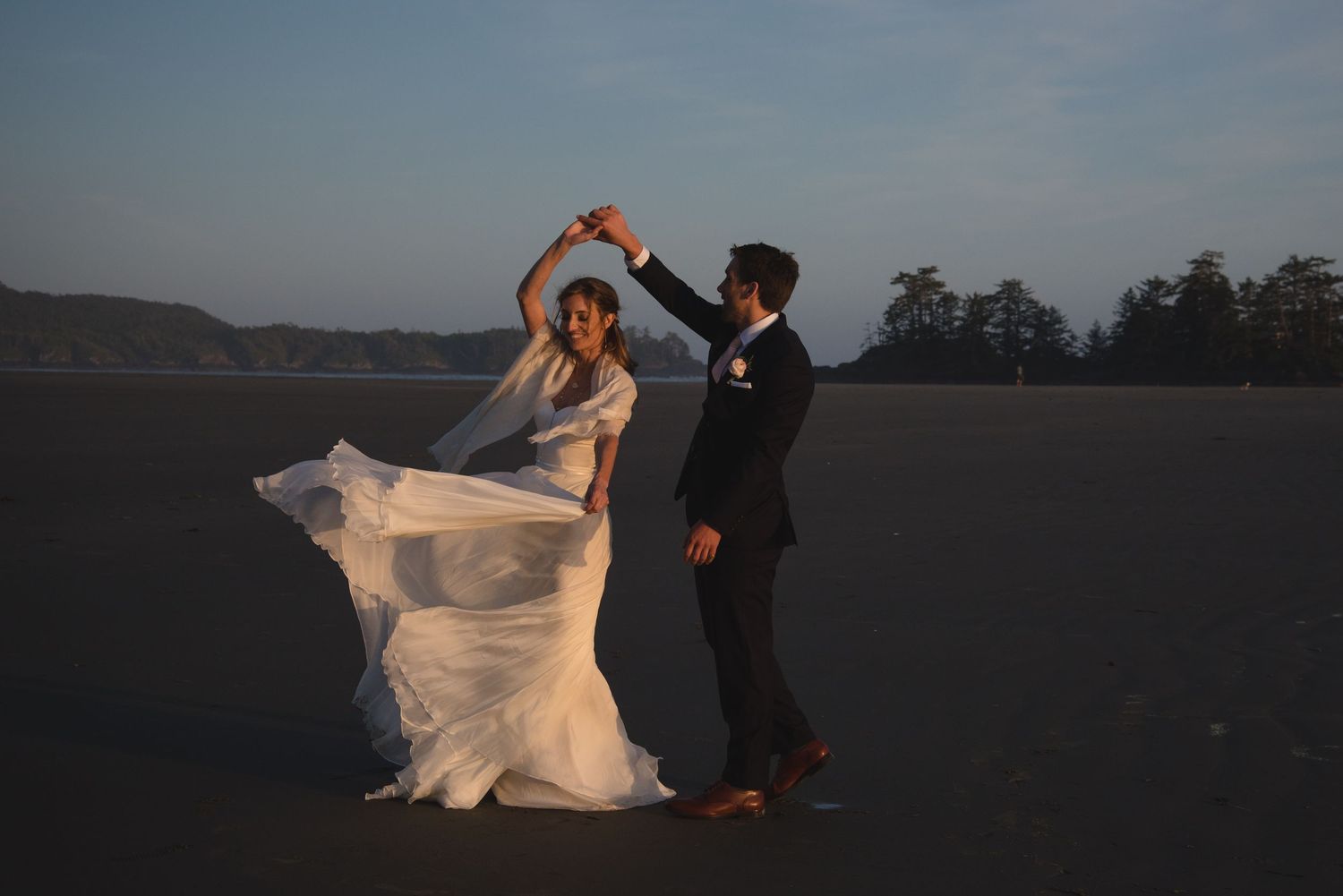A couple dancing together at dusk in an open field as the bride's white dress flows in the wind.