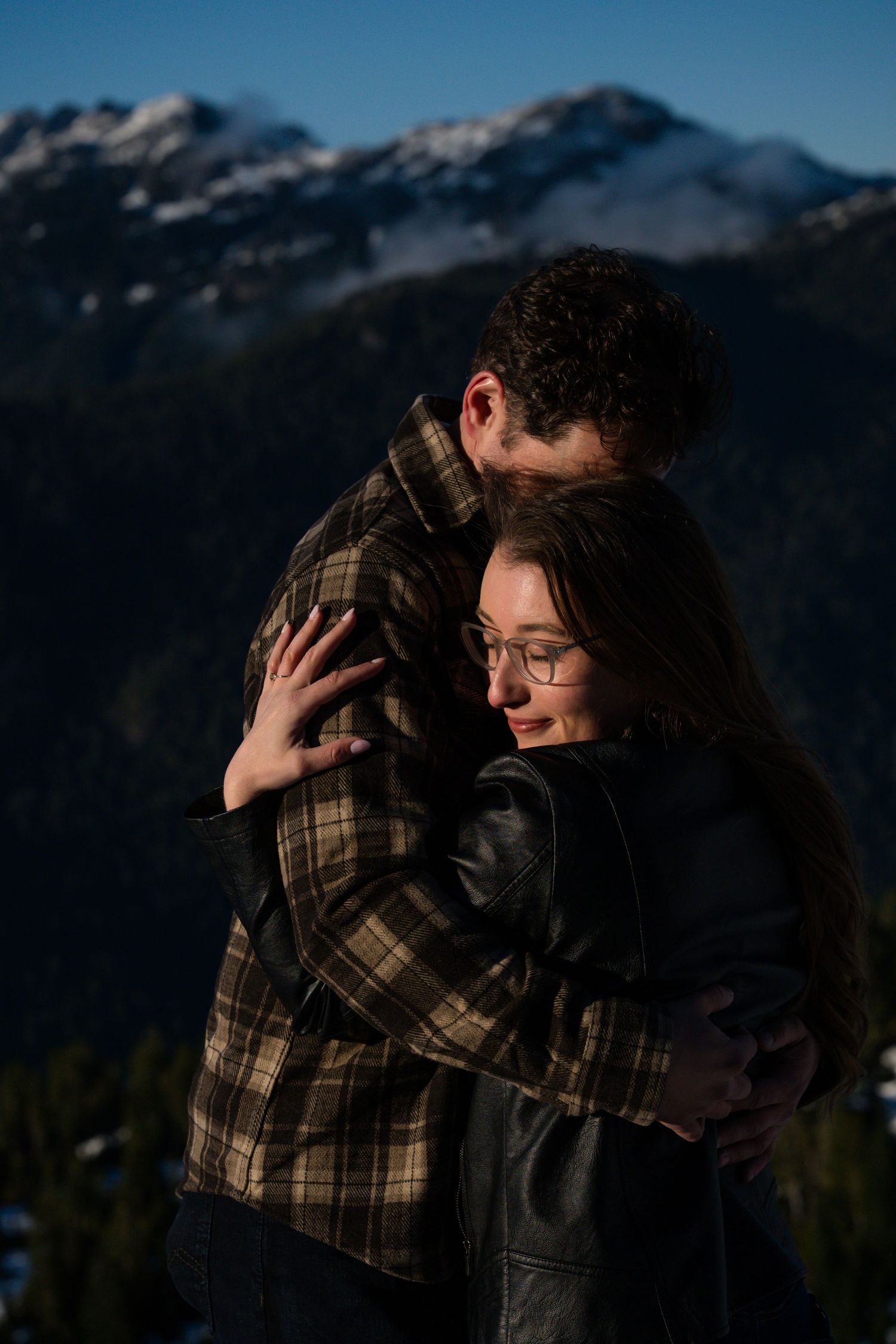 Couple embracing warmly with snow-capped mountains in the background on a sunny day.