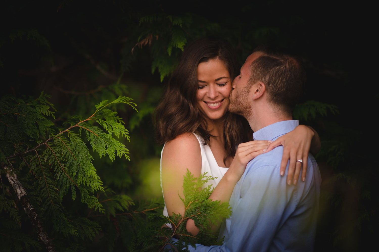 A romantic couple shares an intimate moment in a dark setting with green foliage in the background.