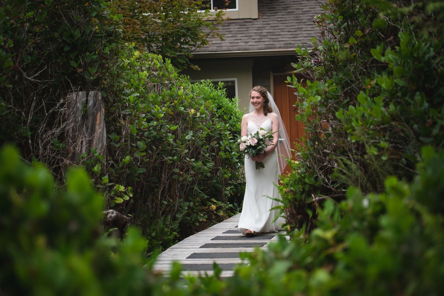 A bride in a white dress holds a bouquet while standing on a garden path surrounded by lush greenery.